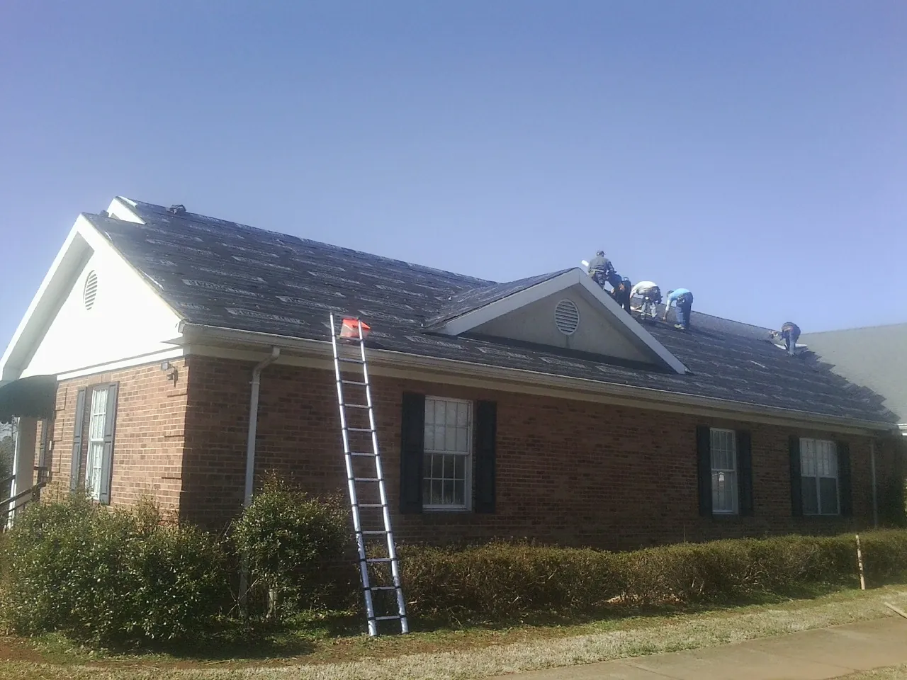 Crew tearing shingles off a brick ranch roof with a ladder set — Emergency Roof Repair work by our crew in Brooten
