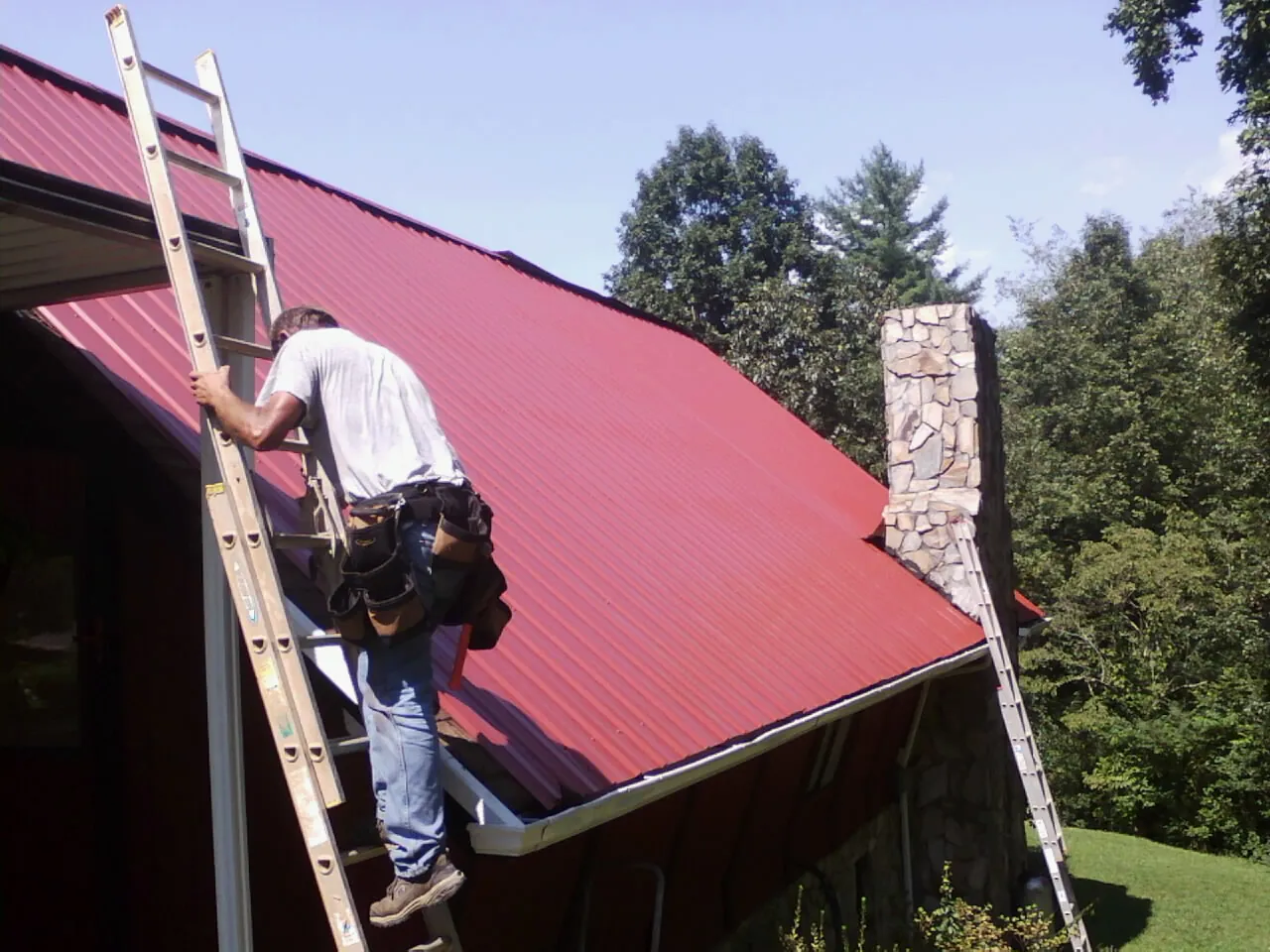 Roofer on a ladder beside a red metal roof and stone chimney — Soffit & Fascia Repair by Roofing Brothers in Brooten